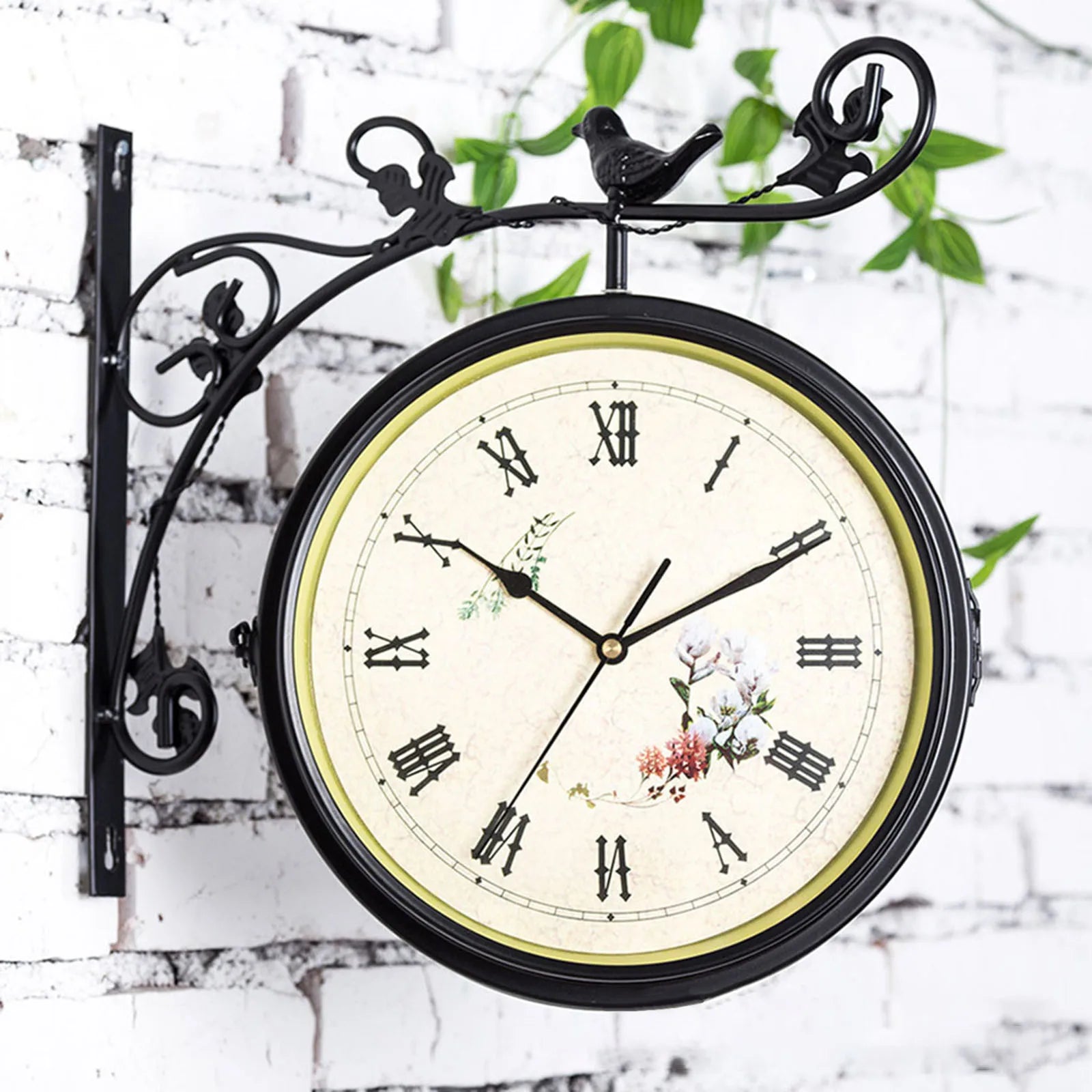 Double sided silent wall mounted clock 25cm diameter with Roman numerals and floral design on a black metal bracket against a white brick wall.