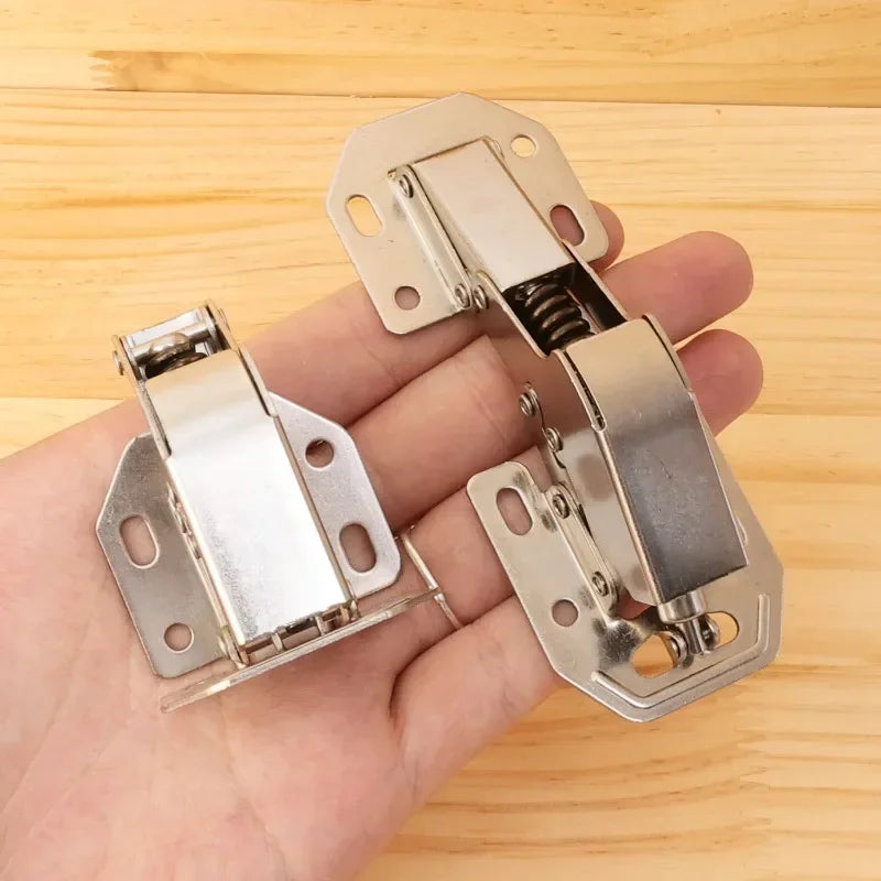 Close-up of two metallic cabinet hinges with hydraulic buffer and soft close feature, displayed on a hand against a wooden background.