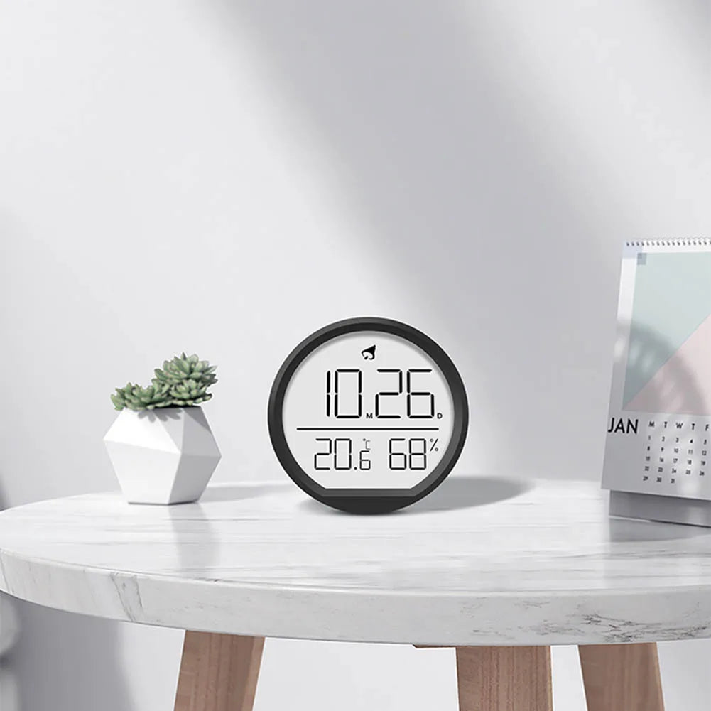 Multi-function electronic clock displaying time, date, temperature, and humidity on a white table with a succulent and calendar.