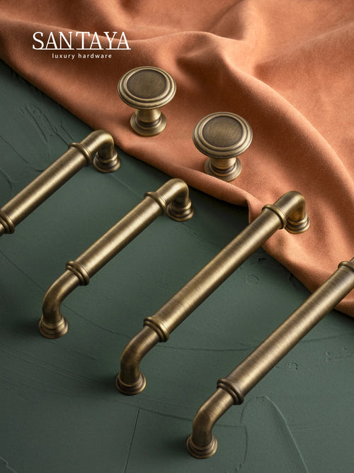 Antique brass cabinet pulls and drawer knobs on textured green surface with orange fabric backdrop.