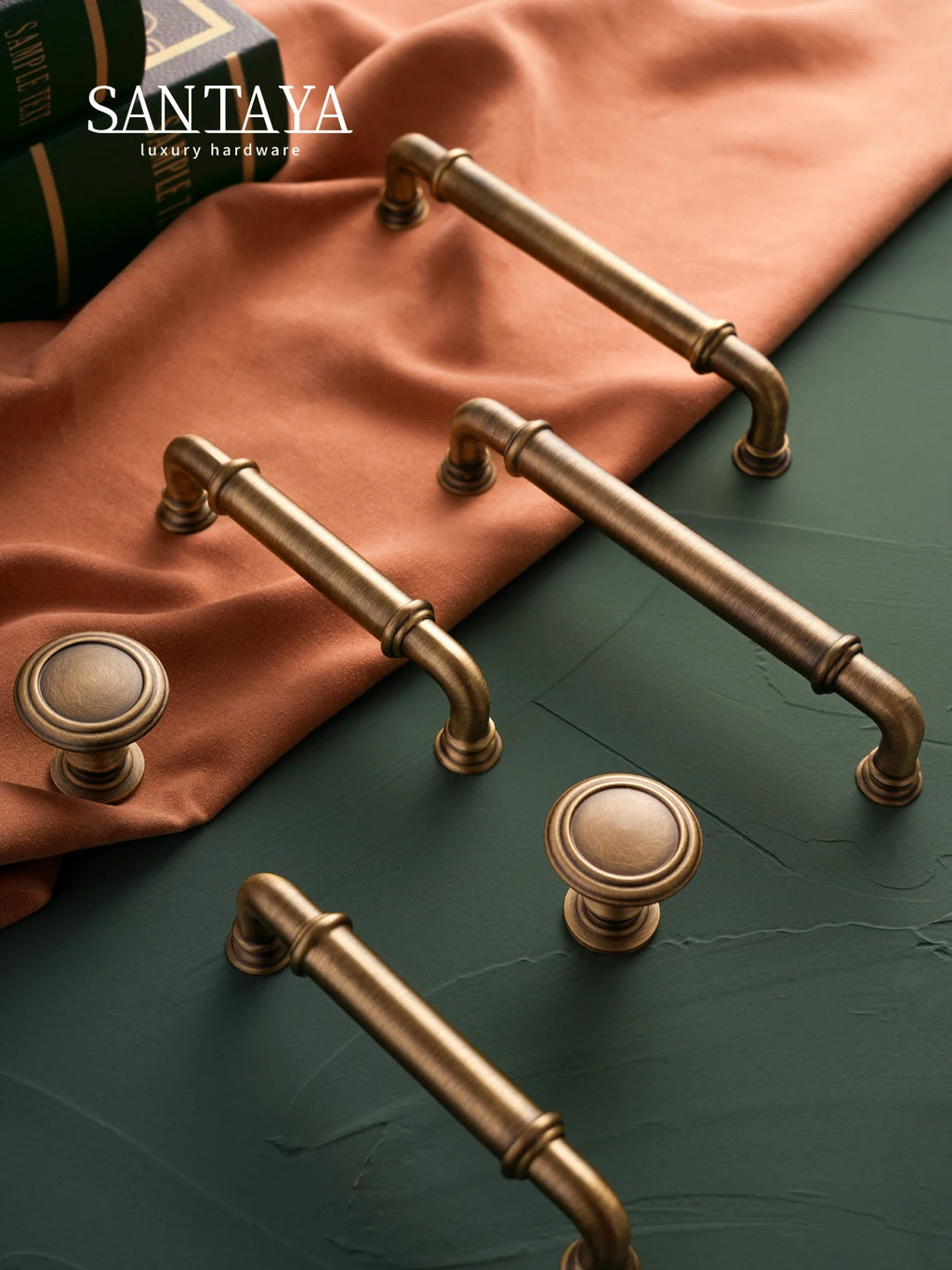 Antique brass cabinet pulls and knobs on green surface with orange fabric, luxury SANTAYA hardware.