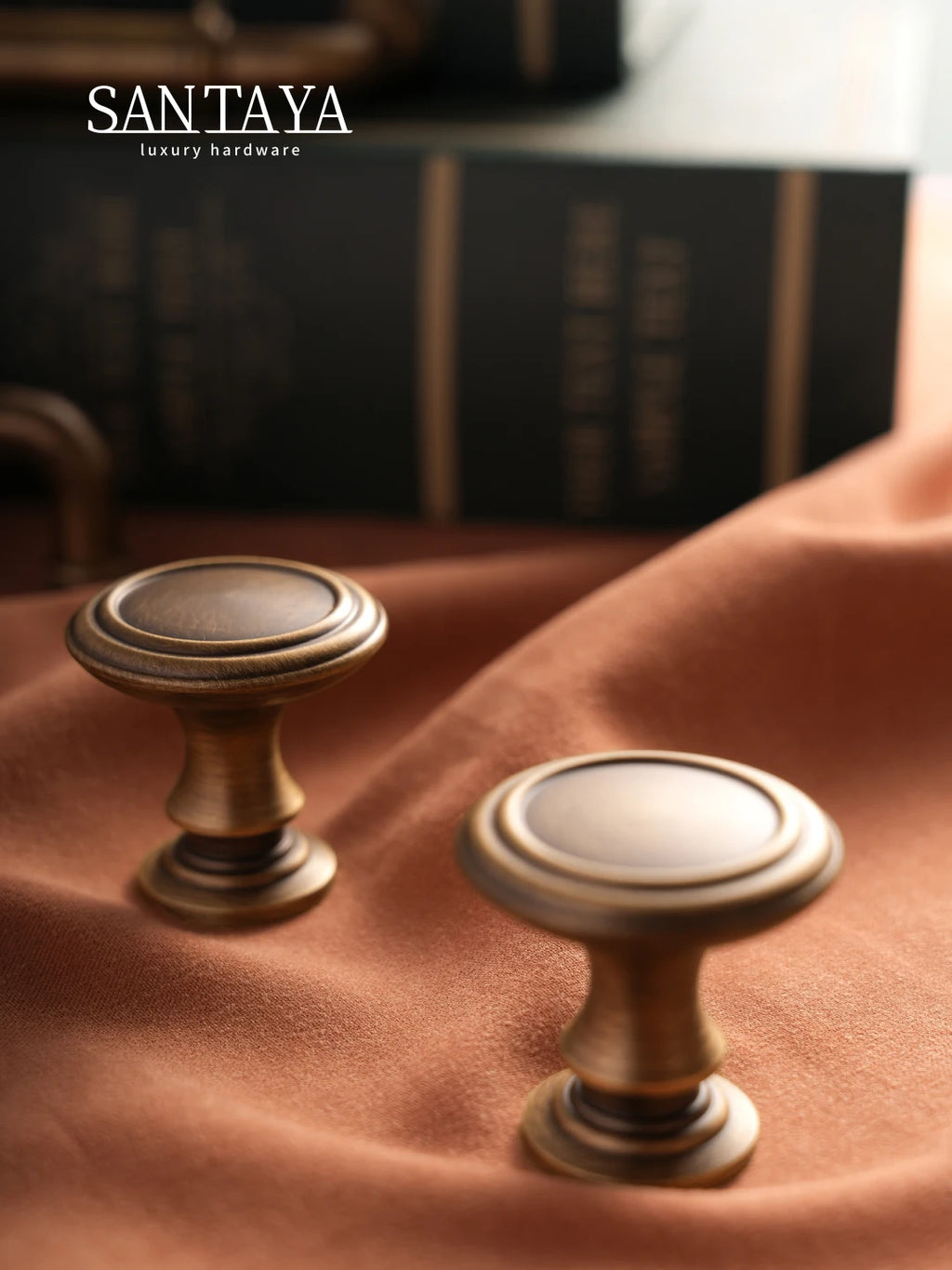 Antique brass cabinet pulls on soft brown fabric with books in the background, showcasing elegant design and detailing.