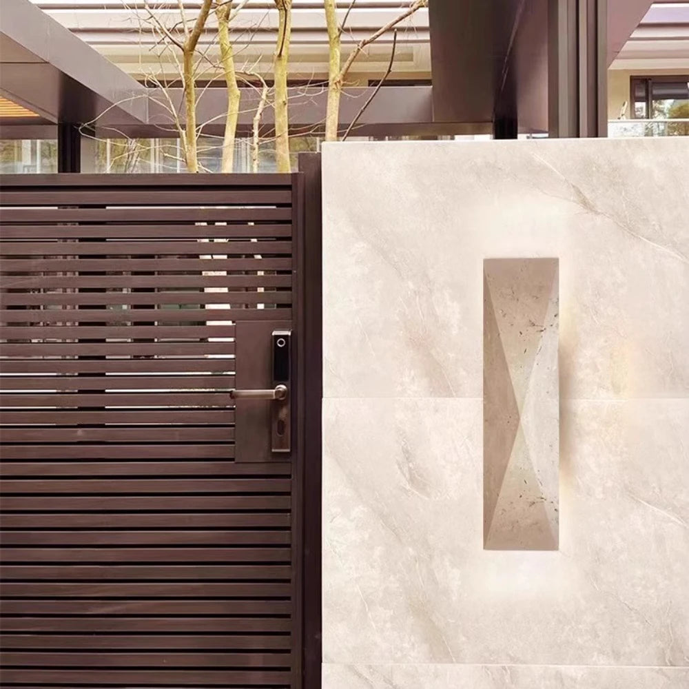 Modern travertine stone wall lamp with geometric design on a light stone wall, next to a dark brown slatted gate in an outdoor courtyard.