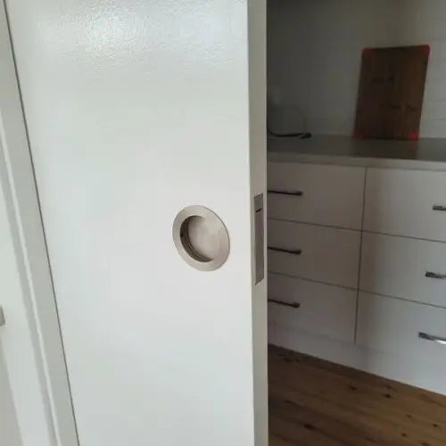 Stainless steel round recessed handle on a white sliding door in a modern kitchen setting.